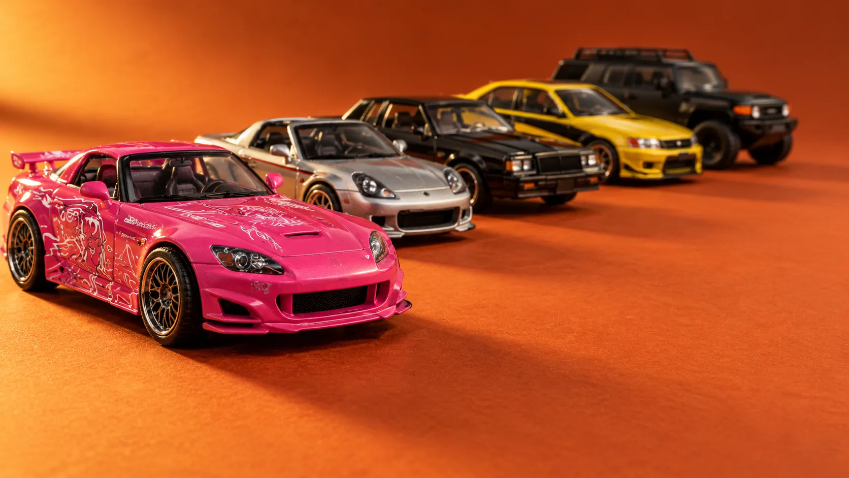 Five die-cast toy cars on orange paper: a pink roadster in front, then a silver coupé, a black muscle saloon, a yellow saloon and a matte black off-roader